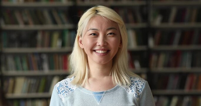 Close up head shot of Asian teenager attractive student girl with dyed blonde hair and wide toothy smile pose look at camera in campus library. University education pupil portrait, studentship concept