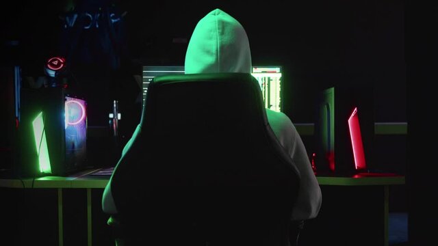 Portrait of a mysterious hacker in a hood, sitting at a table and looking at the camera after hacking a database in a dark room with animation of system codes in the background.