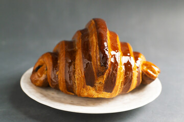Freshly backed french croissant with chocolate filling lies on a white saucer on a gray table....