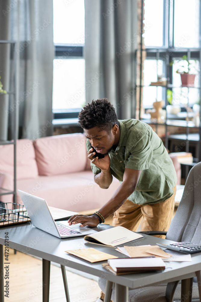 Poster serious young blackman standing at table and checking information on laptop while talking by phone i