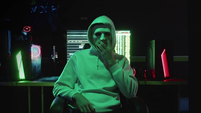 Portrait of a mysterious hacker in a hood, sitting at a table and looking at the camera after hacking a database in a dark room with animation of system codes in the background.