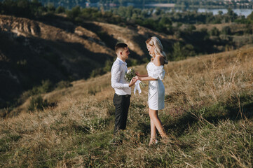 A loving groom and a beautiful blonde bride in a white dress are walking, holding hands, in nature on the background of hills and cliffs in autumn in sunny weather. Wedding photography.