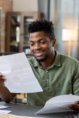 Smiling young Blackman with beard sitting at table in living room and receiving letter from bank