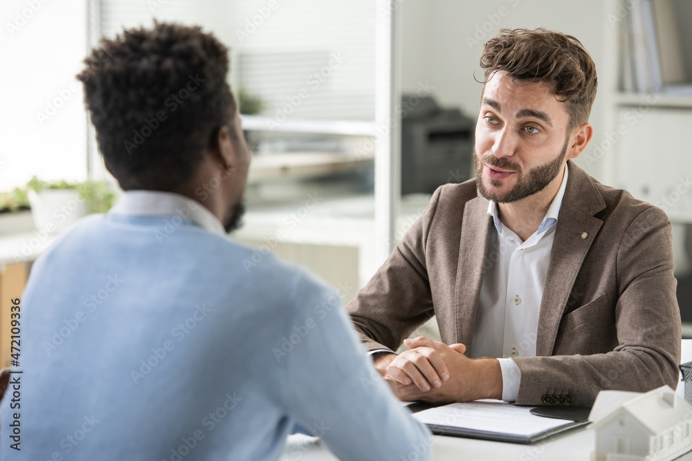 Poster confident young bearded recruiter sitting at table and asking candidate while holding job interview