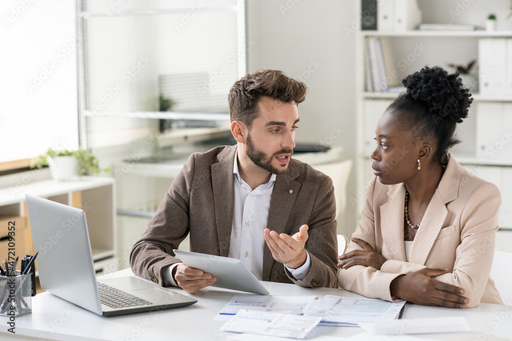 Poster young bearded manager using tablet while discussing income of company with colleague