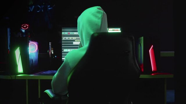 Portrait of a mysterious hacker in a hood, sitting at a table and looking at the camera after hacking a database in a dark room with animation of system codes in the background.