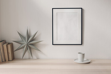Empty black picture frame on white living room wall. Stationery mockup scene with grey folded paper star, cup of coffee and books on wooden table backgroum. Christmas decoration, winter composition.