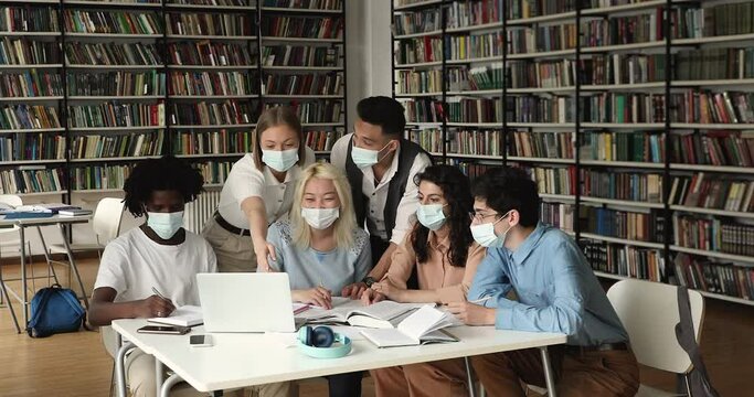 Six diverse multinational highschool students in protective face masks sit at desk in library studying together use laptop. Gen Z, education using modern tech, studies during pandemic outbreak concept
