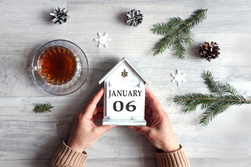 Calendar for January 6: hands hold a decorative house with the name of the month and the numbers 0 and 6, a cup with hot tea, snowflakes, fir branches on a gray background, top view