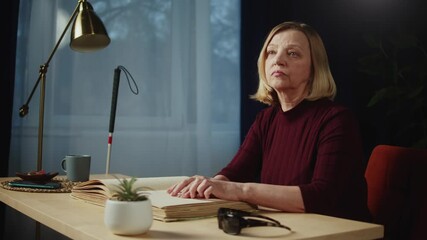 Blind woman reading braille book using fingers, sitting in living room, poorly seeing female person learning to read, home education for people with disabilities, touching letters on sheet of paper.