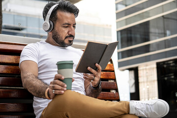 latin man with beard sitting on a bench reading and mug coffee