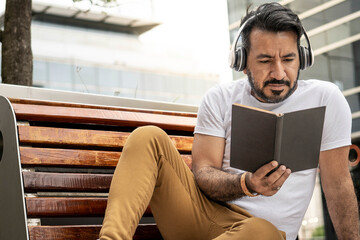 latin man with beard sitting on a bench reading a book in an urban setting