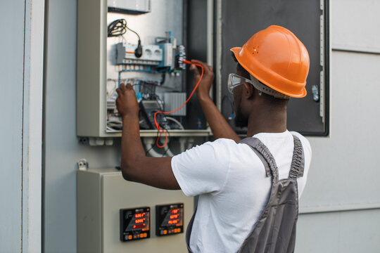 African american repairman in orange helmet and safety glasses checking cables in power transformer among solar station. Competent technician controlling process of green energy production.