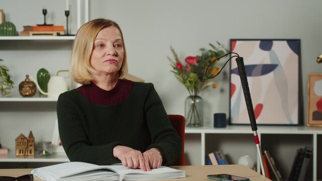 Blind woman reading braille book using fingers, sitting in living room, poorly seeing female person learning to read, home education for people with disabilities, touching letters on sheet of paper.