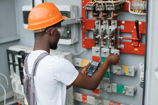 African american electrician in safety helmet and glasses checking voltage in switchgear while working at solar station. Repairing of modern equipment. Production of green energy.