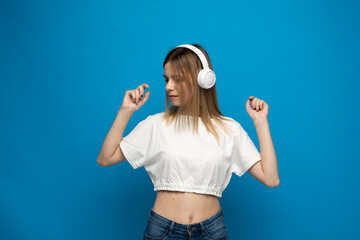 Beautiful young woman in headphones listening to music and dancing on blue background. Young beautiful woman in bright outfit enjoying a music.