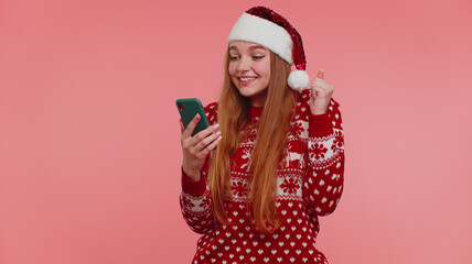 Woman in fashionable Christmas red sweater and hat looking smartphone display sincerely rejoicing win, receiving message with good news, success luck. Girl isolated on pink background. Happy New Year