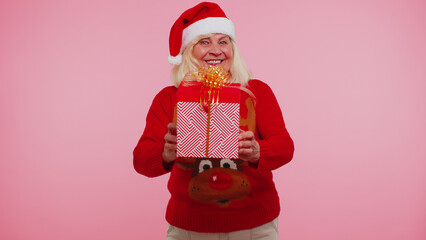 Mature grandmother old woman wears red New Year sweater presenting Christmas gift box stretches out hands isolated on pink studio wall background. Happy Christmas celebration holiday shopping sale eve