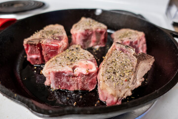 Close up view of several juicy lamb chops sizzling in a hot cast iron skillet on a white stove top