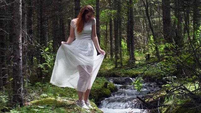 Beautiful happy woman in white dress walks along green forest path on the bank of scenic mountain river. Cheerful young girl in a great mood dancing on the nature background.