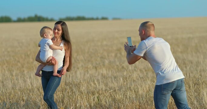Family photoshoot in a wheat field. Father takes pictures of a beautiful mother with a baby daughter on his smartphone, the mother holds the child in her arms and smiles. Family photo for memory. 4k