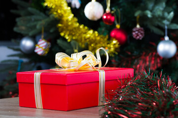 Red gift box with gold ribbon on blurry background with decorated Christmas tree