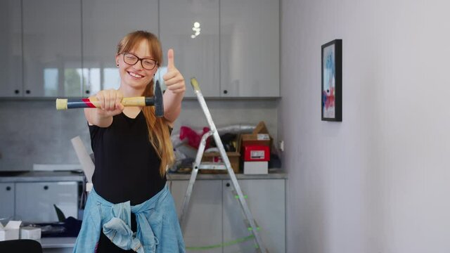 Young crafty independent woman looking into the camera and showing the thumb up while holding the hammer. House renovation and DIY project concept.
