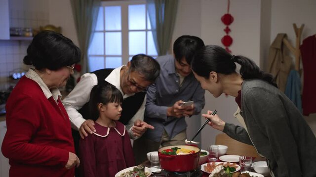 excited asian male father taking photos of family getting together for chinese new year’s dinner by dining table with mobile phone while young mother is cooking hot pot