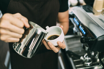 Crop image of a young male barista pouring hot milk into hot espresso black coffee for making Latte Art.