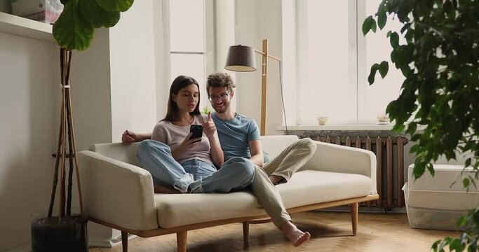 Happy young woman sit on couch lean back to beloved man speak about news at social media show photos on phone screen. Millennial wife rest on sofa share good ideas with smiling husband using telephone