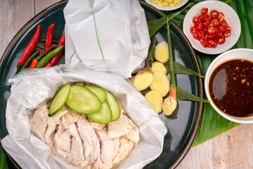 Steamed rice topped with chicken in paper wrap on banana leaves, Hainanese chicken rice served with chilli sauce on a wooden background,