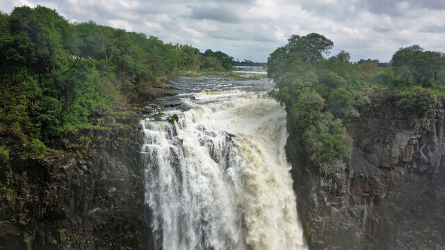 The flow of the Zambezi River collapses into the abyss. Fog over the swirling water. Steep rocky slopes of the gorge, lush vegetation on the plateau. Cloudy sky. Victoria Falls. Zimbabwe © Вера 