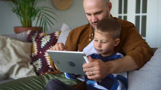 Father and son surf internet. Positive bearded parent teaches curious little boy to use contemporary tablet sitting on sofa in living room closeup spbd