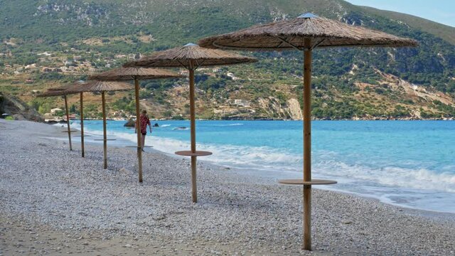 A Male Tourist Relaxing Walking By The Beautiful Shore With Native Umbrellas And Mountains Background At Agia Kiriaki Beach Near Zola Village, Kefalonia, Greece. - Static Shot