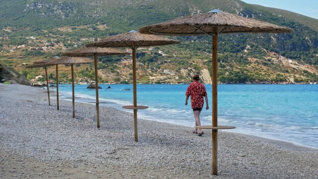 A Man In Summer Beach Outfit Walking Along The Shoreline Pebbled Beach Of Agia Kiriaki, Zola In Kefalonia, Greece. - Wide Shot