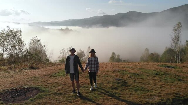 Aerial drone view of young couple walk on meadow with amazing foggy mountains and cloudy background