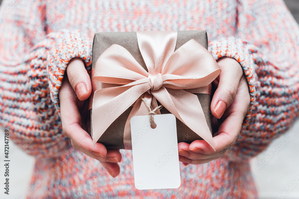 Wall mural Hands holding gift box wrapped in craft paper with perfect pink ribbon and empty tag in cozy knitted sweater. Mockup. Festive concept.