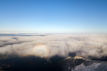 Aerial view from above of white puffy clouds covering snowy mountain tops in bright sunny day