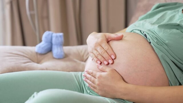 Caring future mother stroking her big tummy while resting on the sofa. Woman caressing her child in the big belly. Two little baby socks are on the armrest at he backdrop.