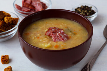 The brown bowl of vegetable soup with meat and crackers. Split pea soup with ham, carrots and potatoes with spoon on a rustic white background. Classic Dutch pea soup erwtensoep, snert with bacon.