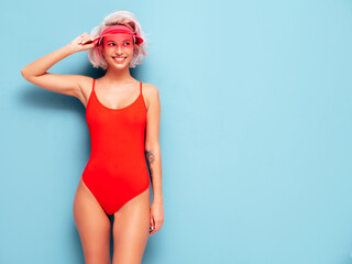 Portrait of young smiling blond model in summer swimwear red bathing suit and transparent visor cap. Sexy carefree woman having fun and going crazy. Female posing near blue wall in studio