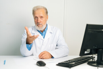 Portrait of serious mature adult male doctor in white uniform coat speaking, looking at camera, gesturing sitting at desk with computer in medical office, consulting patient.