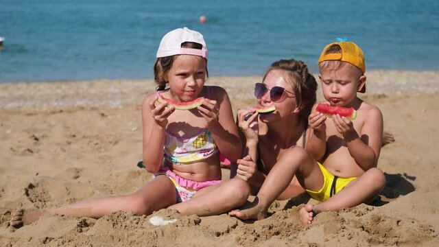 Mom and children are sitting on the beach near the sea and eating watermelon. Vacation. Family in sunglasses and caps relaxing on the beach.