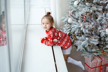 Cute little girl is leaning on the windowsill of a decorated Christmas tree dreamily looking into...