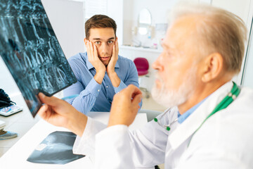 Close-up of mature adult male physician consulting scared young man patient giving bad news explaining results of MRI image. Unhappy frustrated young man listening to bad news, looking at camera.