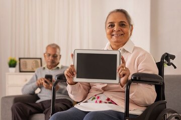 Fototapeta na wymiar Old woman showing digital tablet on wheel chair and old man using smartphone on sofa
