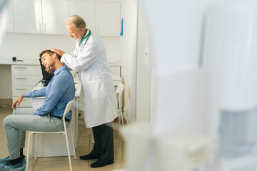 Wide shot of senior adult male manual therapist turning and bending head of male patient to check injured neck. Young man on preventive examination of doctor chiropractor on neck diagnostic.