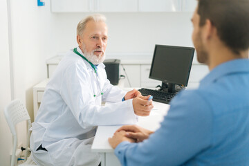 Close-up view from back of unrecognizable male patient communicating with mature adult doctor sitting at table in medical office. Senior physician interviewing sick young man and writing prescription.