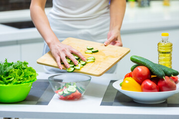 Close up of a girl hands slicing vegetables with a knife on a cutting board for a vegan vitamin vegetable salad and putting them in a glass bowl while cooking breakfast in the kitchen