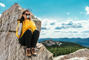 A female tourist is resting after a hard climb up the mountain. A tourist drinks tea from a thermos...